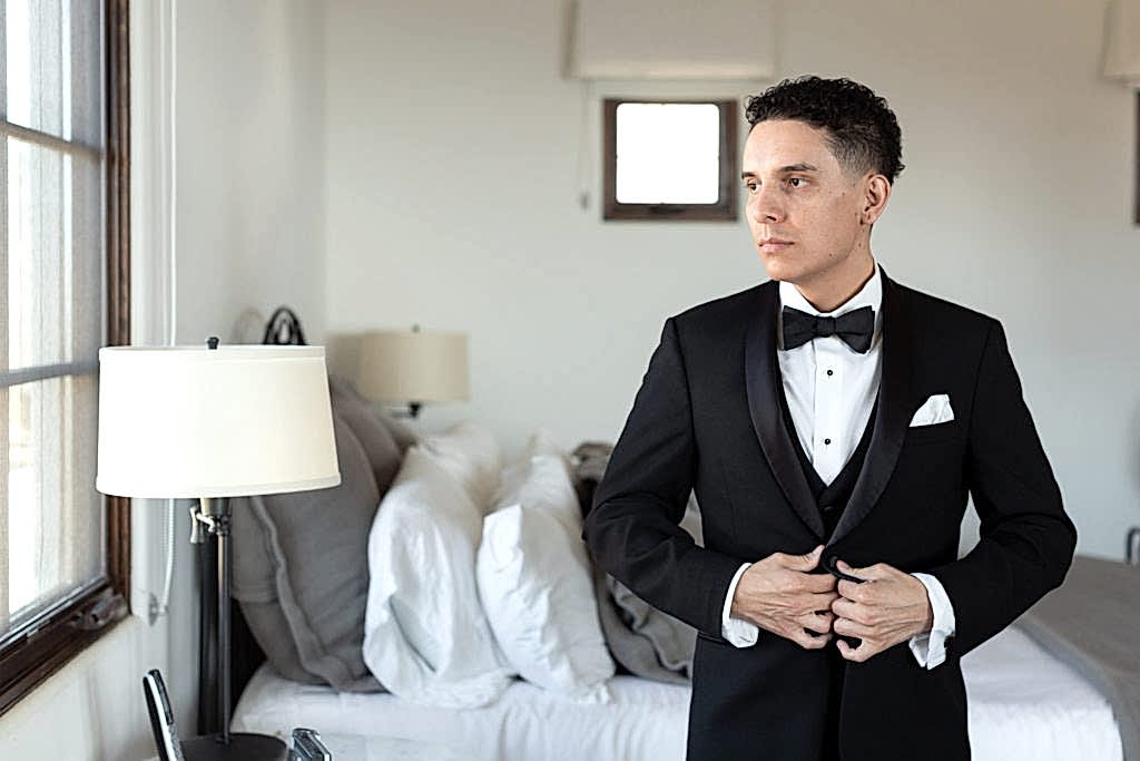 groom adjusting his black tuxedo as he looks out a window as he gets ready for his wedding day