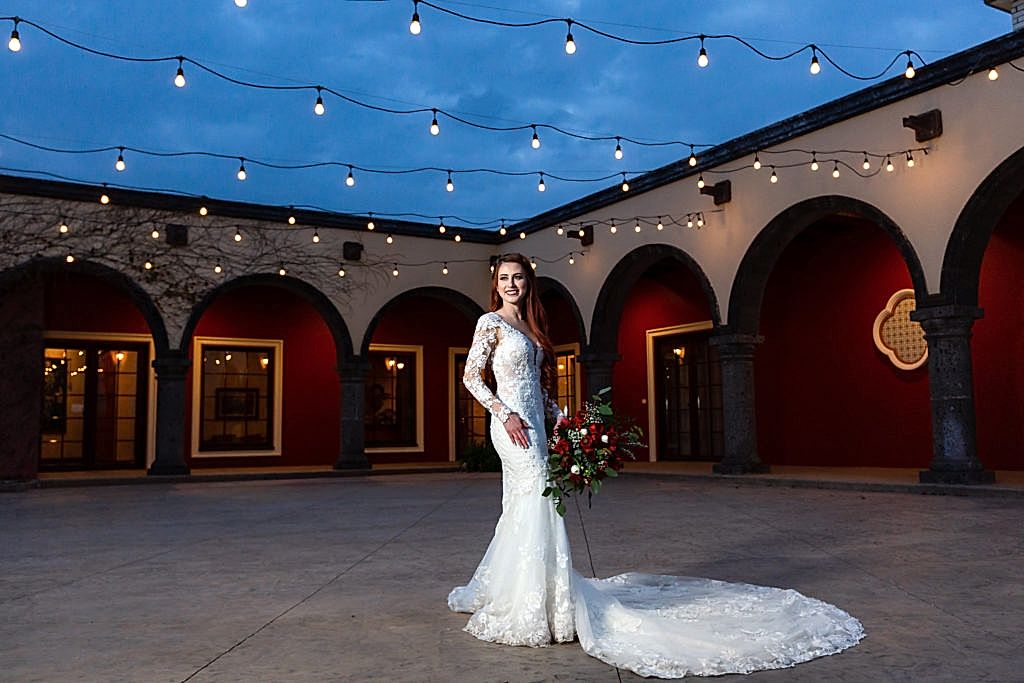 outdoor bridal pictures at night with bride in a lace wedding dress with a large train standing on an outdoor patio with string lights over head