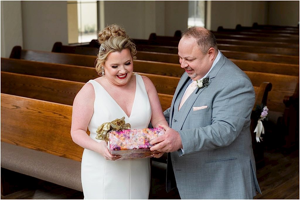 Dallas wedding photographers show bride and groom stand in the Ana Villa chapel holding a wrapped gift with a large gold bow