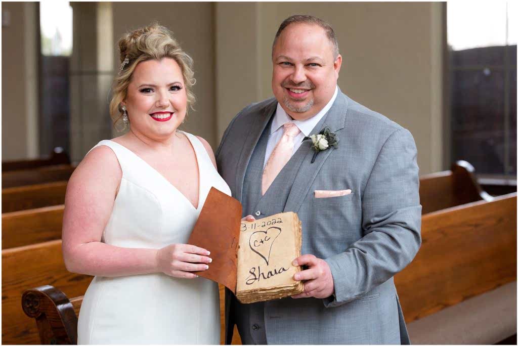 Bride and groom stand side by side displaying a leather and rough paper guestbook with a heart on the front page captured by Dallas wedding photographers
