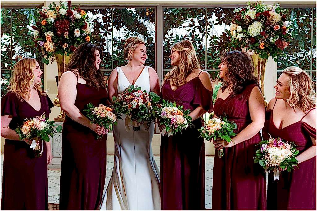 Bride laughs with five bridesmaids in front of floral arrangements at Ana Villa TX chapel by Dallas wedding photographers