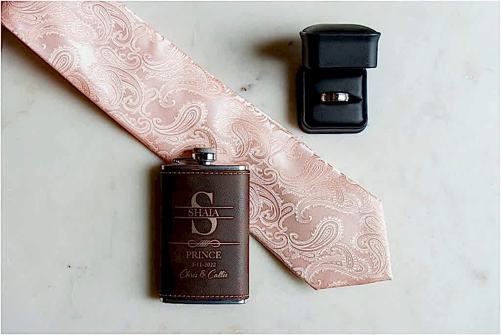 A flatlay of a flask, paisley pink tie, and the groom's ring in a ring box captured by Dallas wedding photographers
