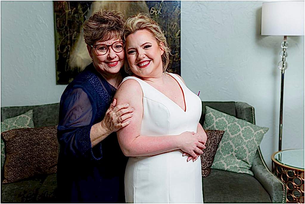 Dallas wedding photographers capture the bride's mother and the bride posing and smiling together in front of a green velvet sofa at The Colony TX