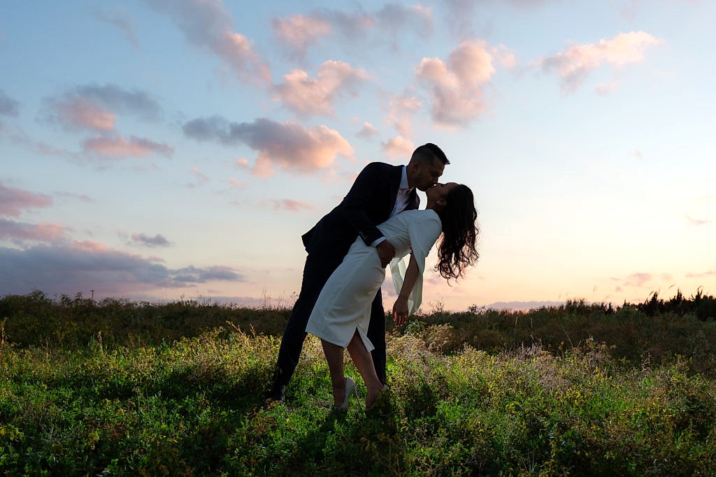 Silhouette photo of couple dipping and kissing on grassy hill with cotton candy sky behind them during a Plano park engagement session