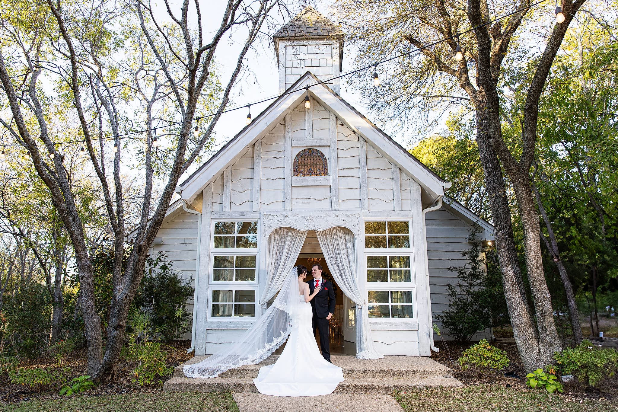 Bride with flying veil and groom standing in doorway of Chandler Chapel at Bella Woods wedding venue in Midlothian TX surrounded by trees