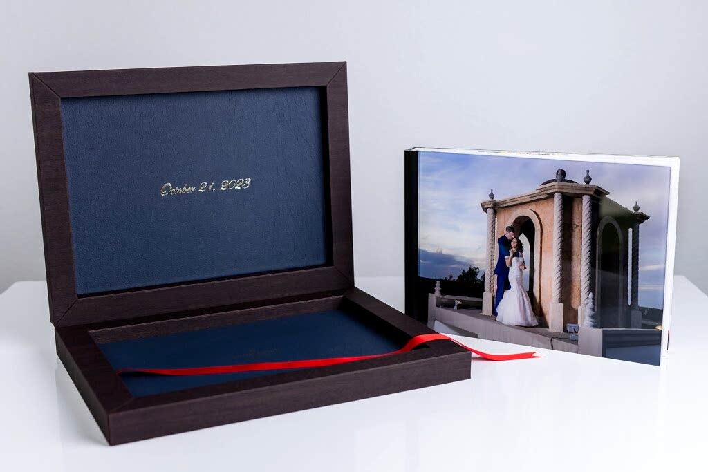 an heirloom wedding album with black spine and an acrylic photo cover of bride and groom hugging on stoney ridge villa wedding venue's rooftop sitting next to it's open luxury dark wood and blue leather box with gold foil and a red ribbon