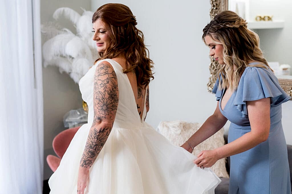 The morning of the wedding bride gets into her wedding dress as her maid of honor who is in a dusty blue bridesmaid dress helps her get ready