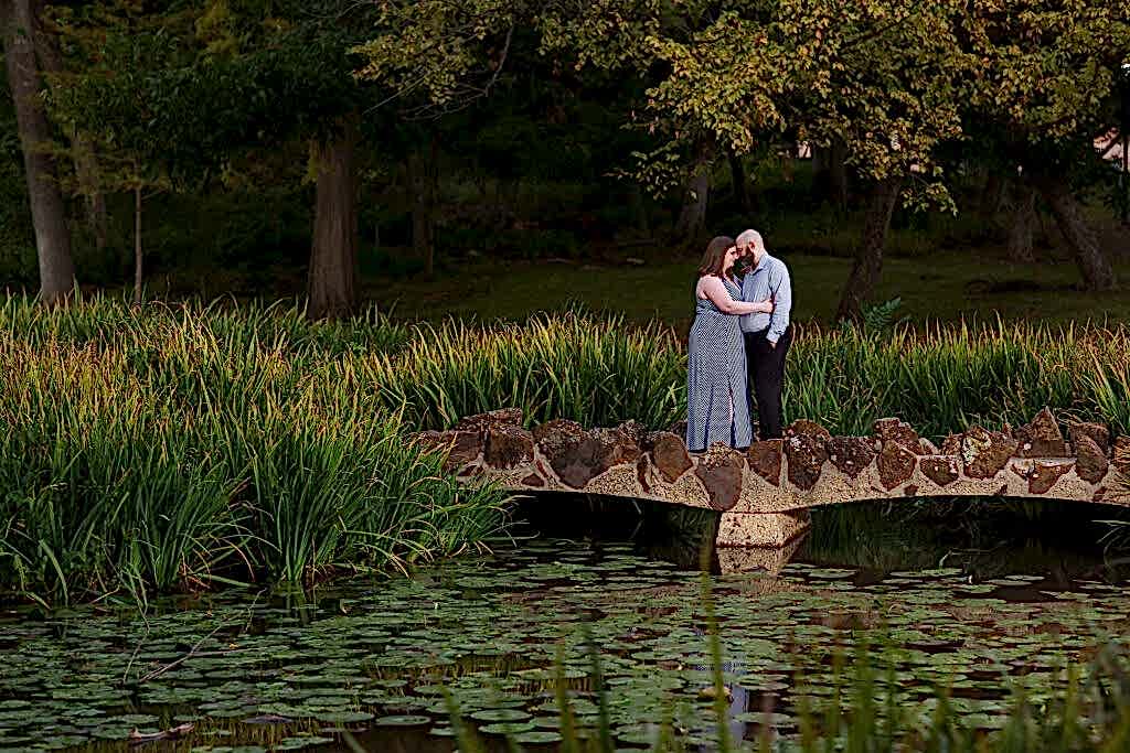 Dallas wedding photographer captures engagement pictures at Texas Women's University with woman leaning onto a man as they stand together on a bridge over a lake filled with Lilly pads for engagement session