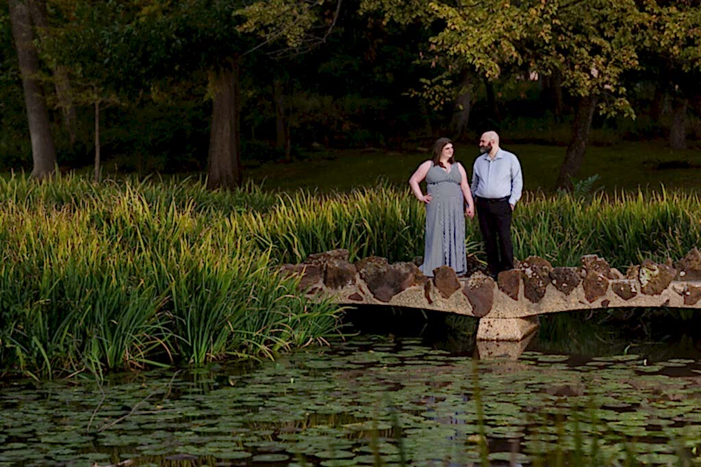 Dallas wedding photographer Stefani Ciotti Photography captures engagement photo at Texas Women's University with engaged couple standing together looking at each other on a bridge over a lake filled with Lilly pads with trees surrounding them