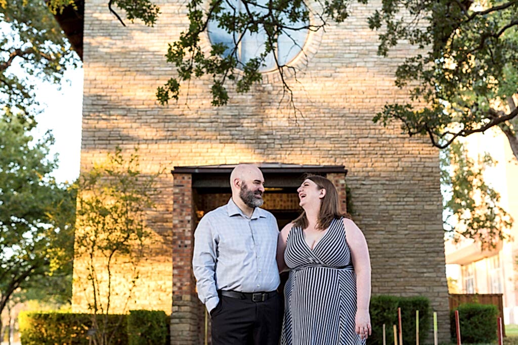 Dallas engagement picture of a man and woman in grey outfits laughing at each other and standing in front of an old building with a round window at Texas Woman's University