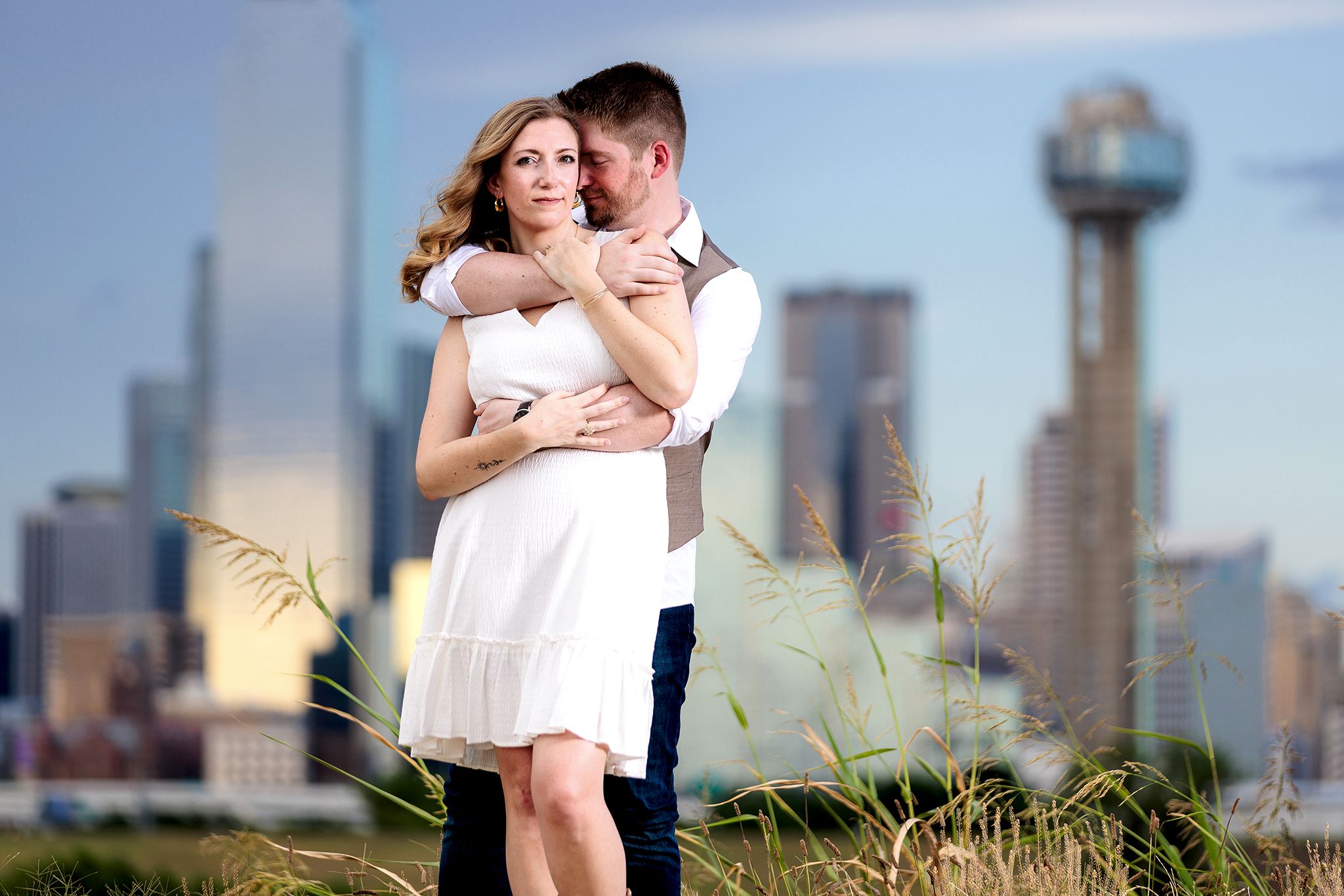 Man nuzzling into his fiancee's neck during outdoor engagement session wearing white dress with downtown Dallas in the background