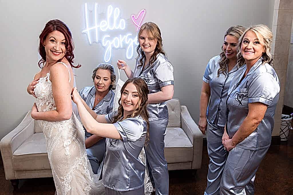 Bridesmaids in wedding pjs laughing and helping bride into wedding dress in bridal suite