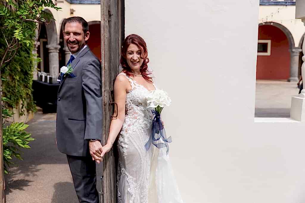 Bride and groom holding hands behind old wooden door for first touch while bride holds white tulip bouquet