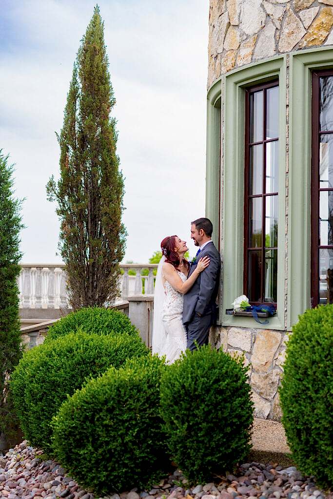 Bride and groom smiling at each other up against wedding venue surrounded by trees and bushes