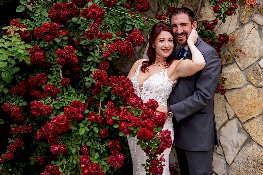 Bride posing with groom behind red roses at Azle TX wedding venue, Stoney Ridge Villa