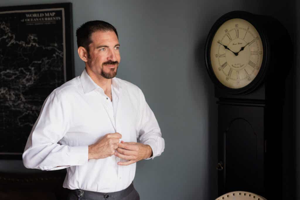 Groom buttoning up dress shirt with old clock in the background in groom's suite