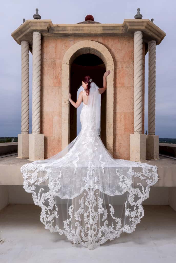 Red-haired bride posing on bell tower on Stoney Ridge Villa wedding venue's rooftop in Azle TX with intricate and laced wedding train flowing