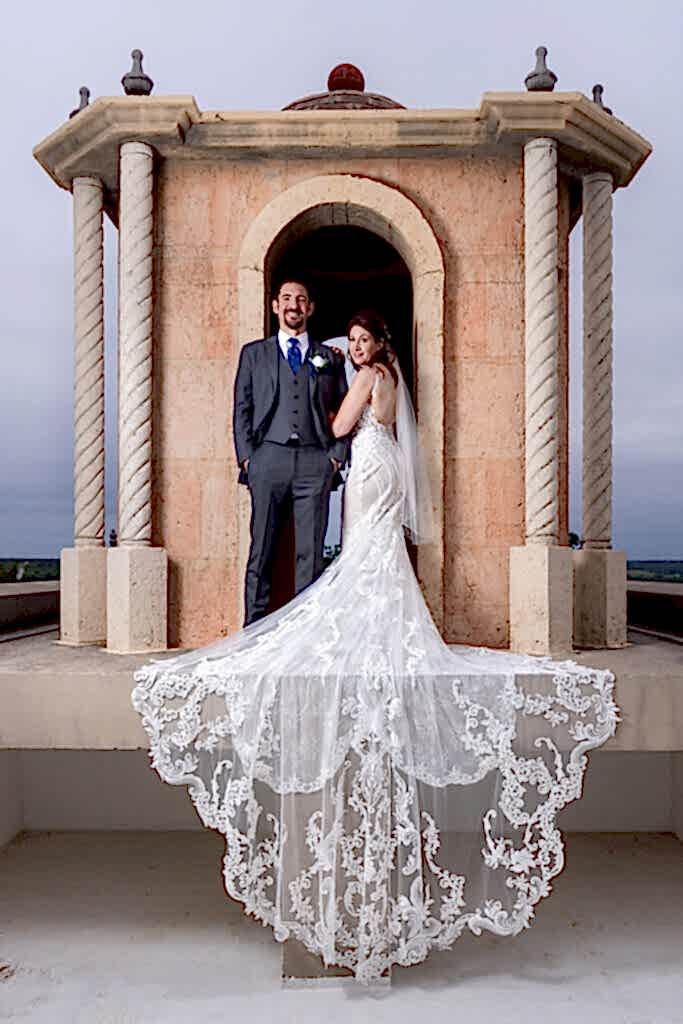 Bride and groom smiling at camera on bell tower on Stoney Ridge Villa wedding venue's rooftop in Azle TX with intricate and laced wedding train flowing