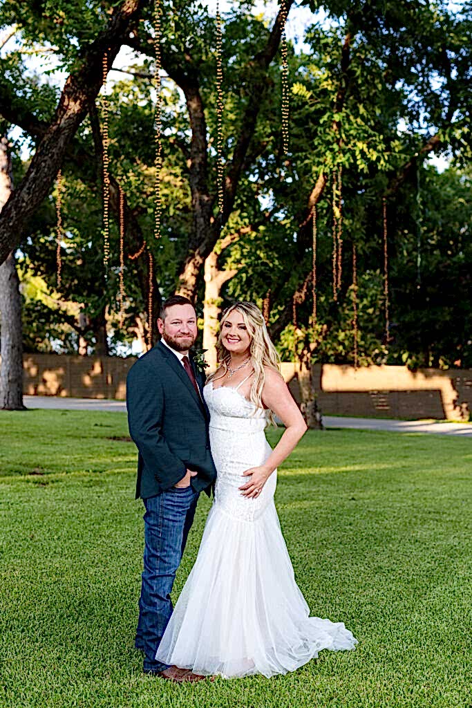 Wedding couple smiling under large lush trees with hanging string lights