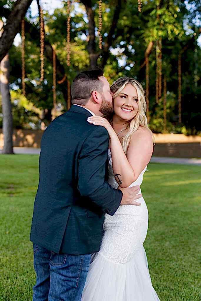 Groom kissing smiling bride on temple under trees and hanging string lights