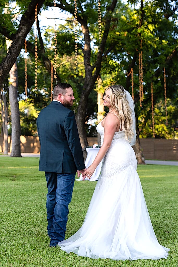 Groom holding brides hands while both looking back over their shoulders smiling at each other under lush trees and hanging string lights