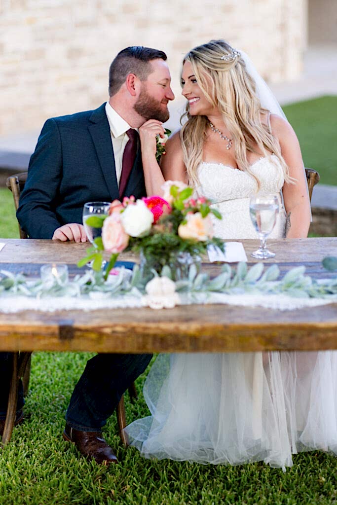 Bride and groom sitting at a rustic sweetheart table smiling closely at each other