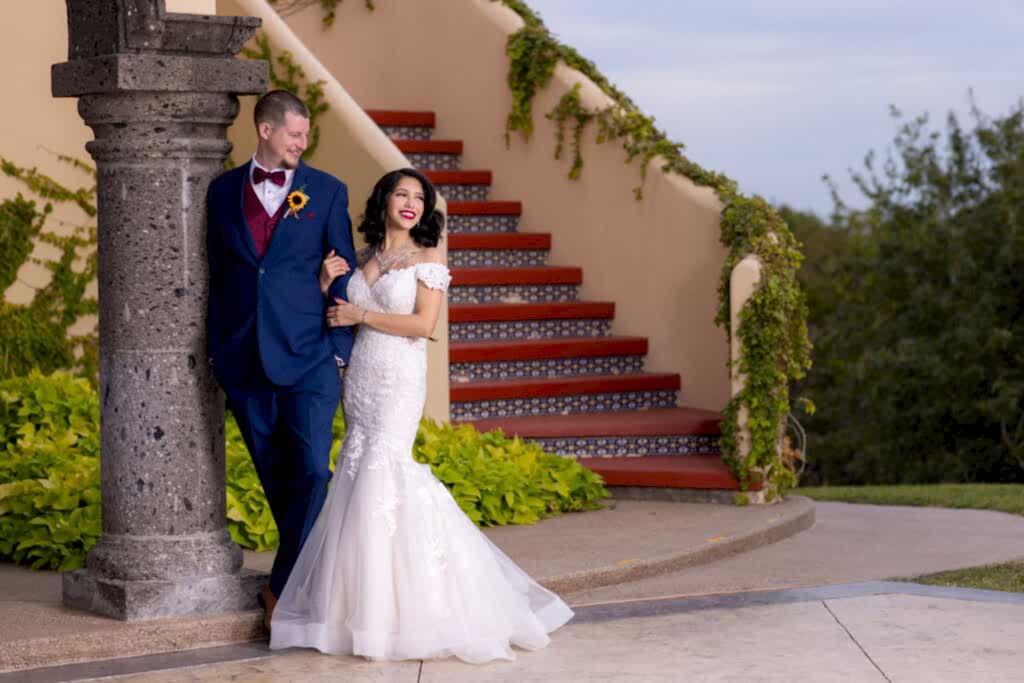 Bride hugging onto groom while he leans on a pillar looking off in front of a staircase at Fort Worth Estate Wedding venue Stoney Ridge Villa
