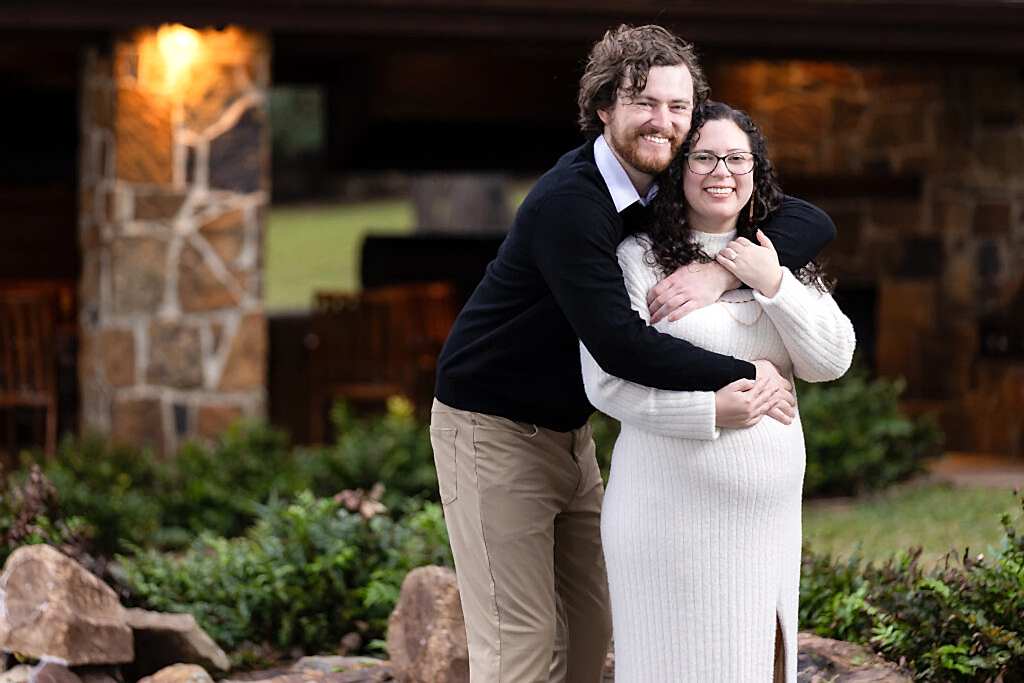 Engaged man hugging fiancée from behind and smiling at camera during Mabank lakehouse engagement session