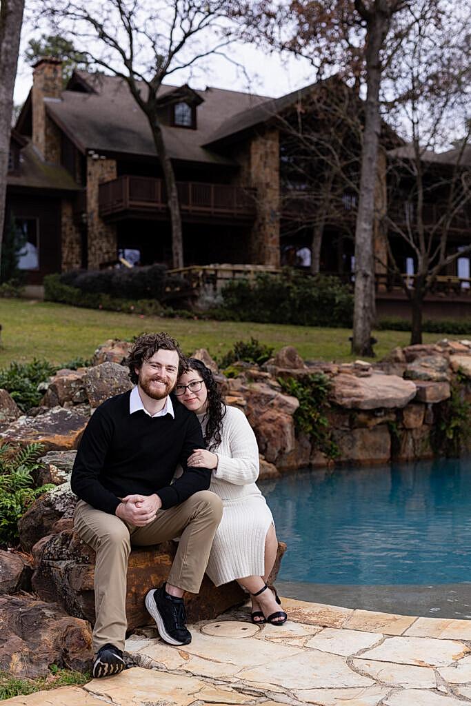 Engaged couple sitting on rocks by pool with their Mabank lakehouse in the background for their engagement session