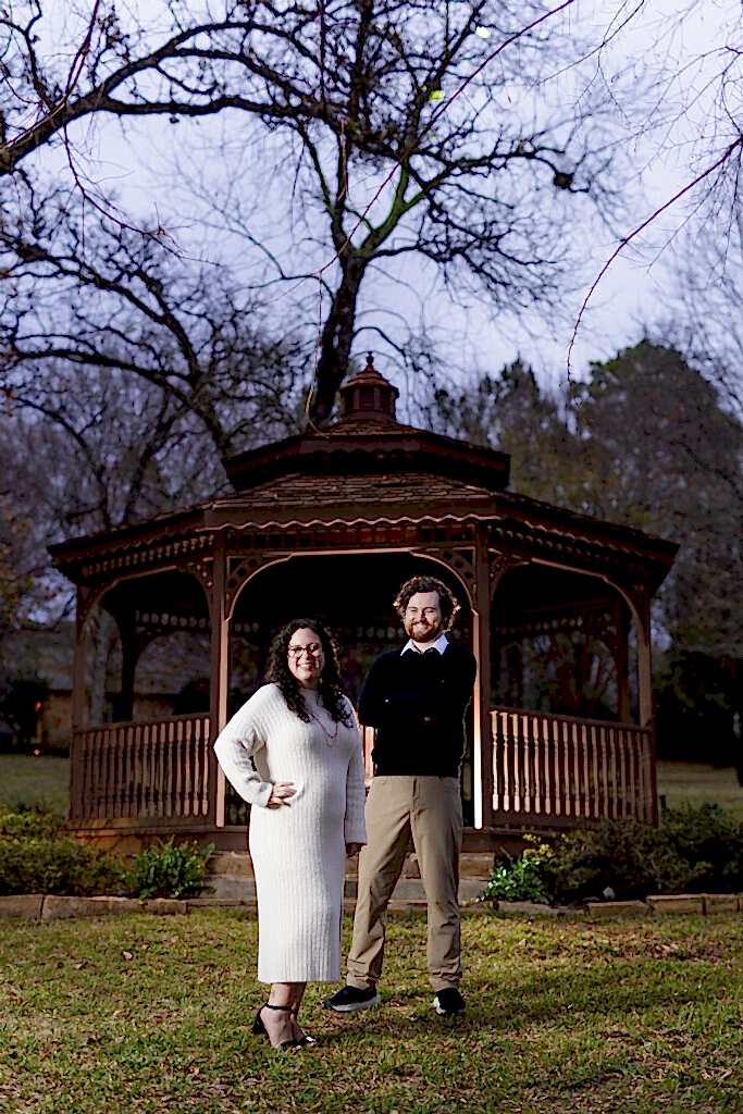 Engaged couple standing and smiling in front of gazebo at night on lake house property with woods in the background during engagement session in North Texas