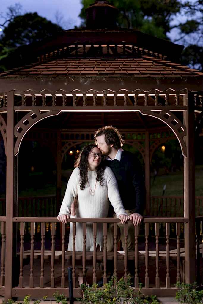 Man kissing fiancée's forehead underneath brown gazebo at nighttime during engagement session