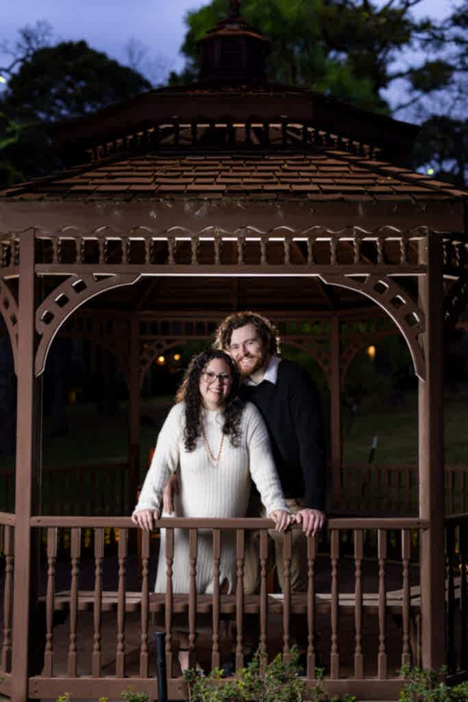 Engaged man and woman standing together underneath brown gazebo at nighttime during engagement session in Mabank TX