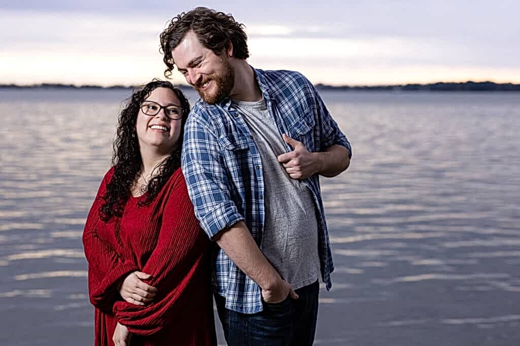 Engaged couple laughing towards each other with lake and sunset behind them during Mabank lakehouse engagement session
