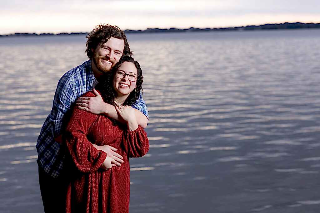 Engaged couple hugging and smiling with lake and sunset behind them during Mabank lakehouse engagement session