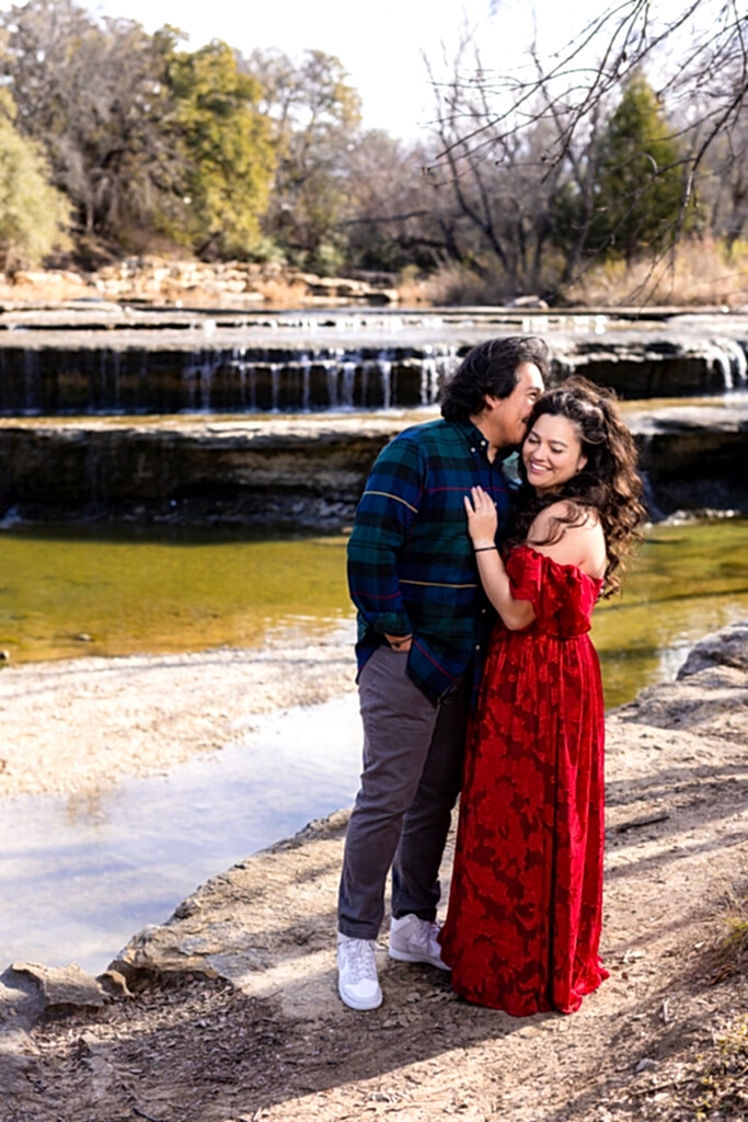 Man kissing fiancée wearing red dress laughing with Airfield Falls waterfall in the background during engagement session in Fort Worth