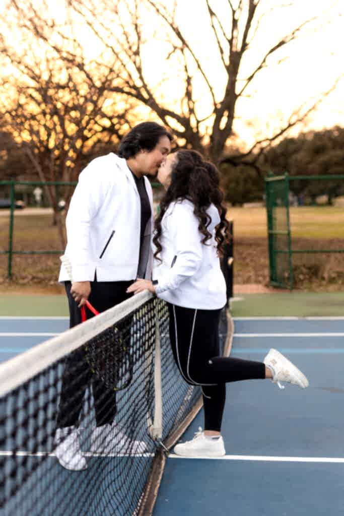 Engaged couple in matching black and white track suits kissing over tennis net during engagement session