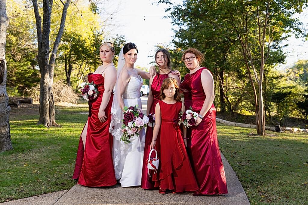 Bride and bridesmaids wearing red surrounded by trees at Bella Woods wedding and event venue in Midlothian Texas