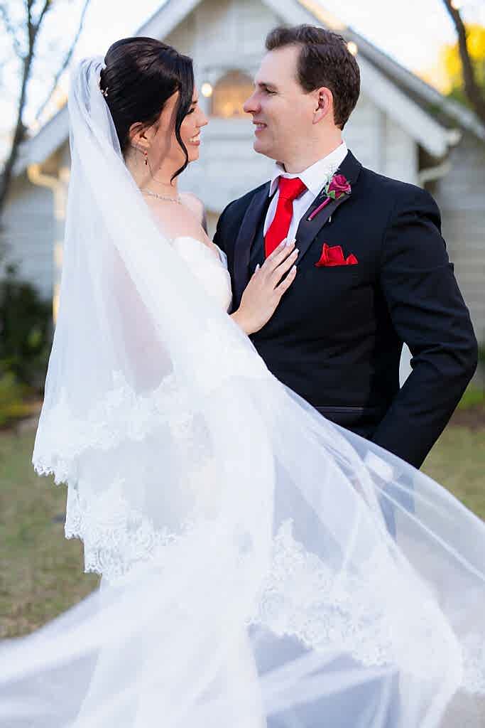 Bride with flying veil and groom standing together and smiling in front of Chandler Chapel at Bella Woods wedding venue