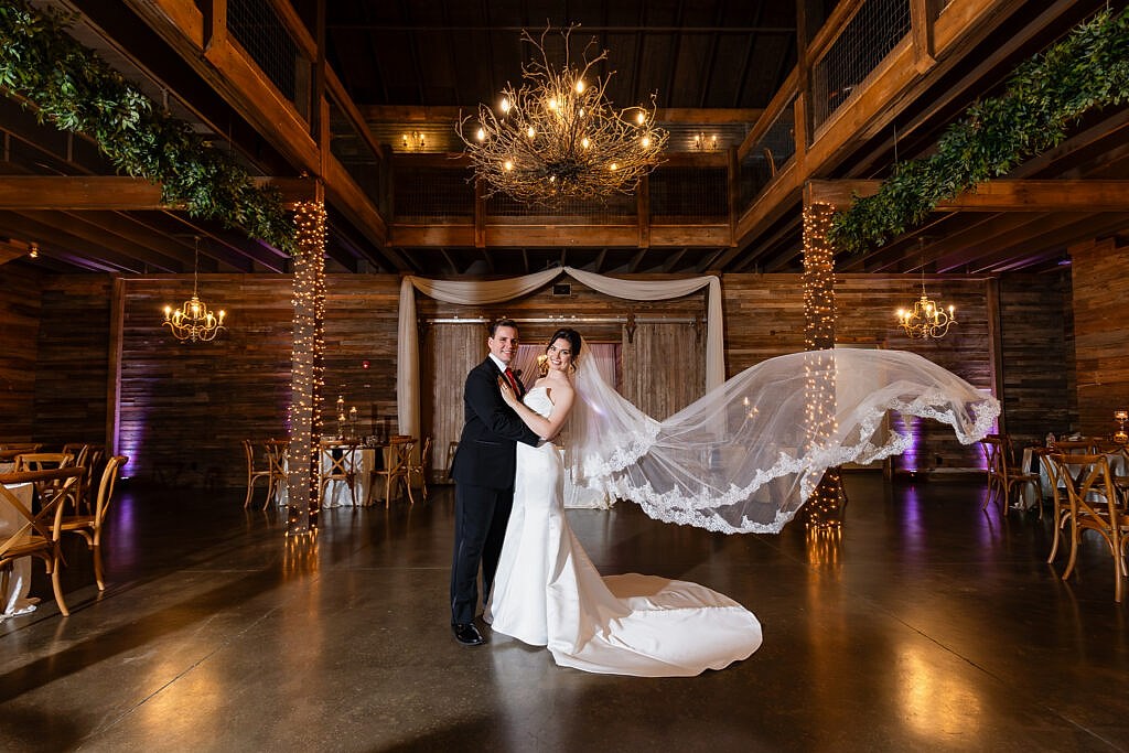 Bride with flying veil and groom smiling inside gorgeous rustic Taylor Barn at Bella Woods wedding venue in Midlothian Texas
