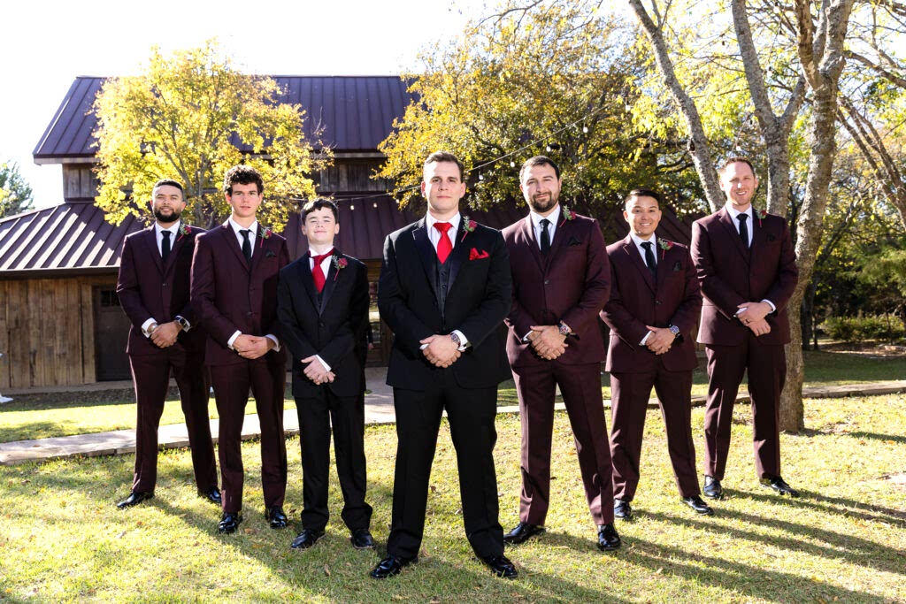 Groom and groomsmen standing in V formation wearing black and maroon suits in front of Taylor Barn at Bella Woods wedding venue
